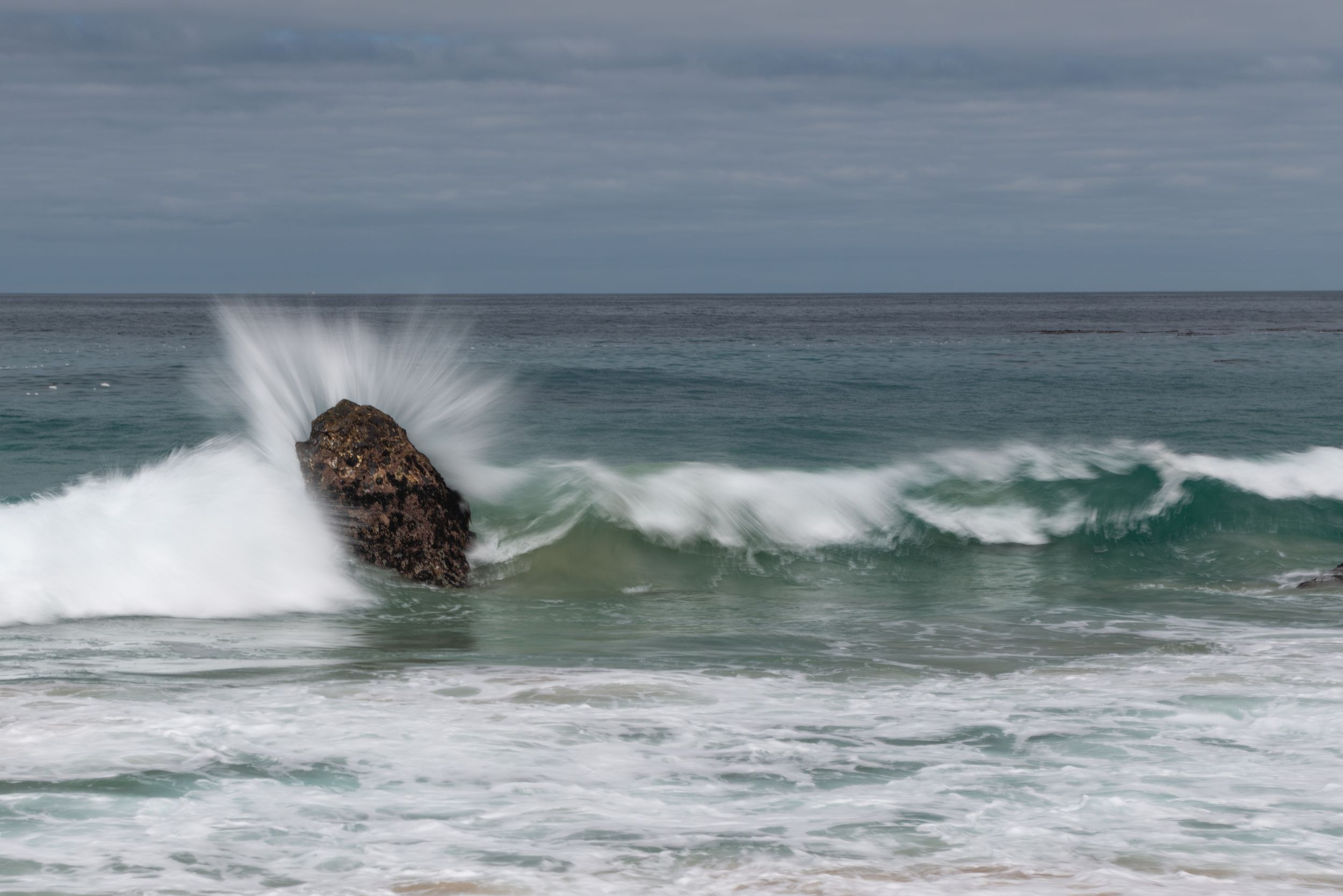 Ocean wave crashing against a dark rock with white sea spray against a turquoise sea and cloudy sky.