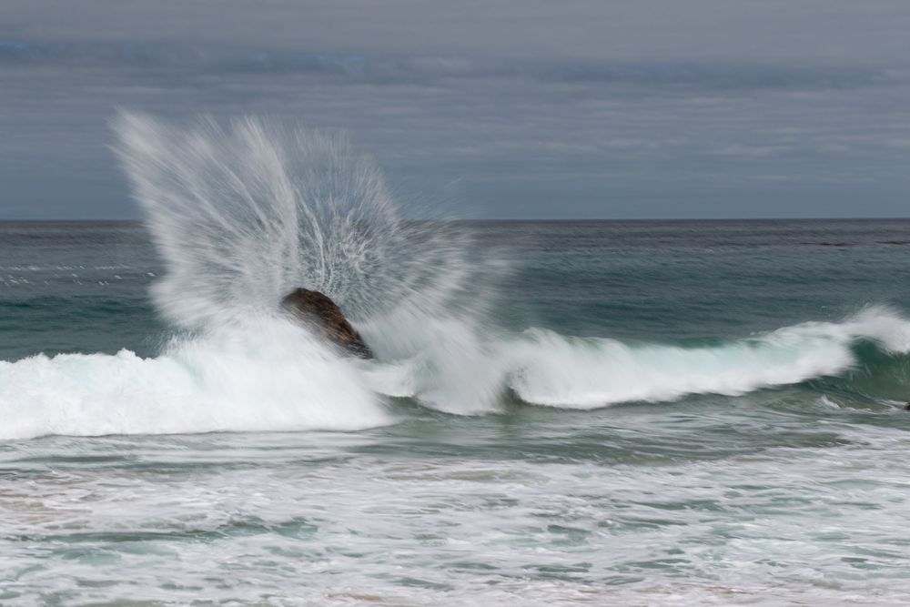 Ocean wave crashing dramatically over a dark rock, sending white sea spray into the air against a cloudy sky.