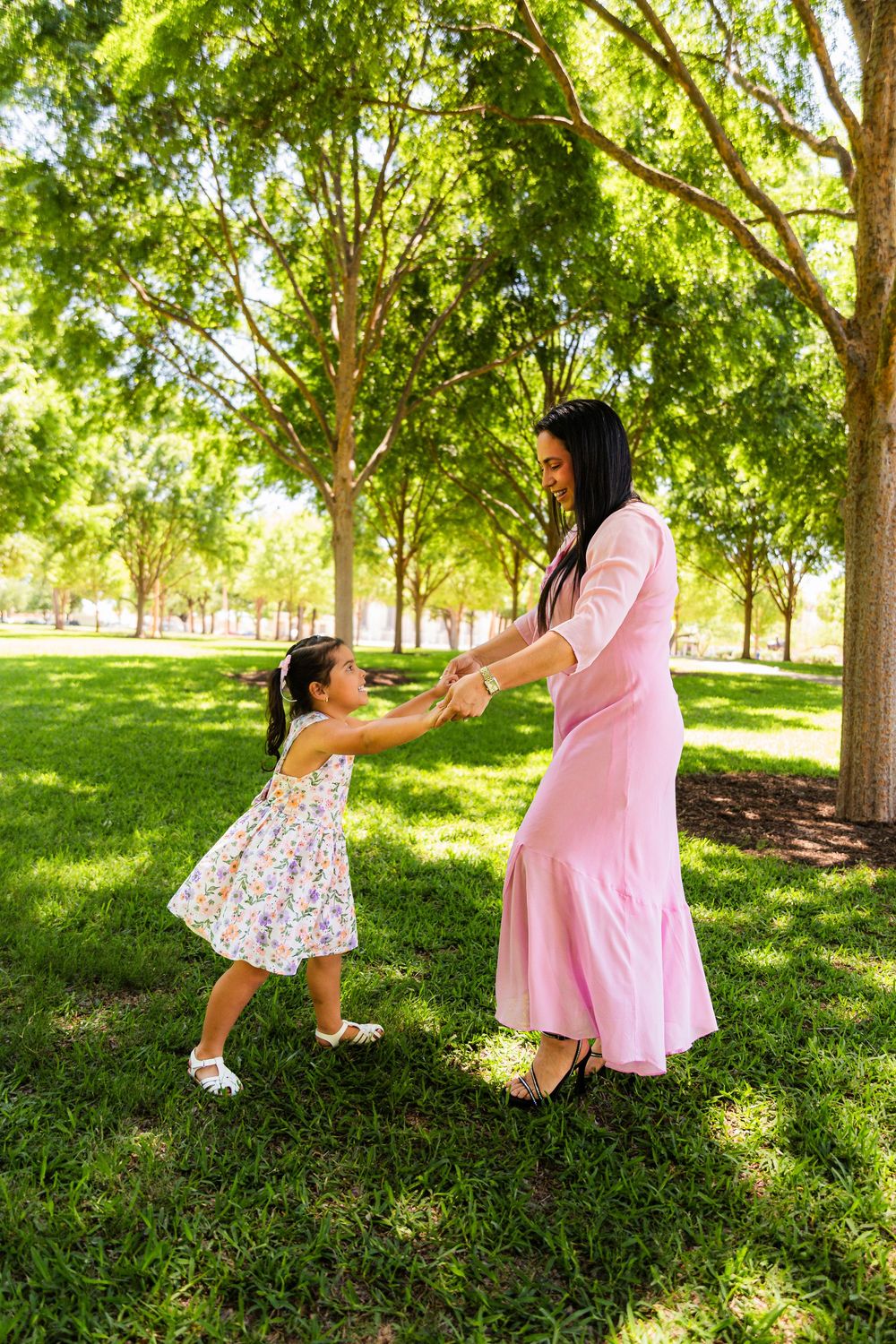 A mother in a pink dress holds hands with her young daughter in a floral dress, dancing together in a sunny green park.