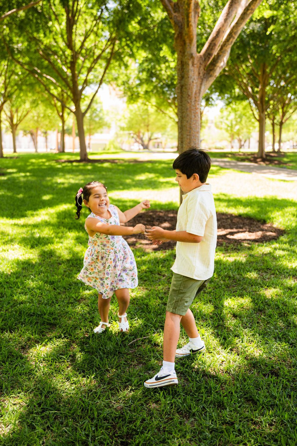 Two children holding hands and dancing together on green grass in a sunny park surrounded by trees.
