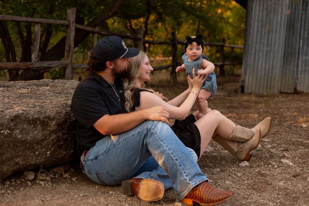 A young couple sits against a rustic barn wall outdoors, lovingly interacting with their baby girl in a charming family portrait.
