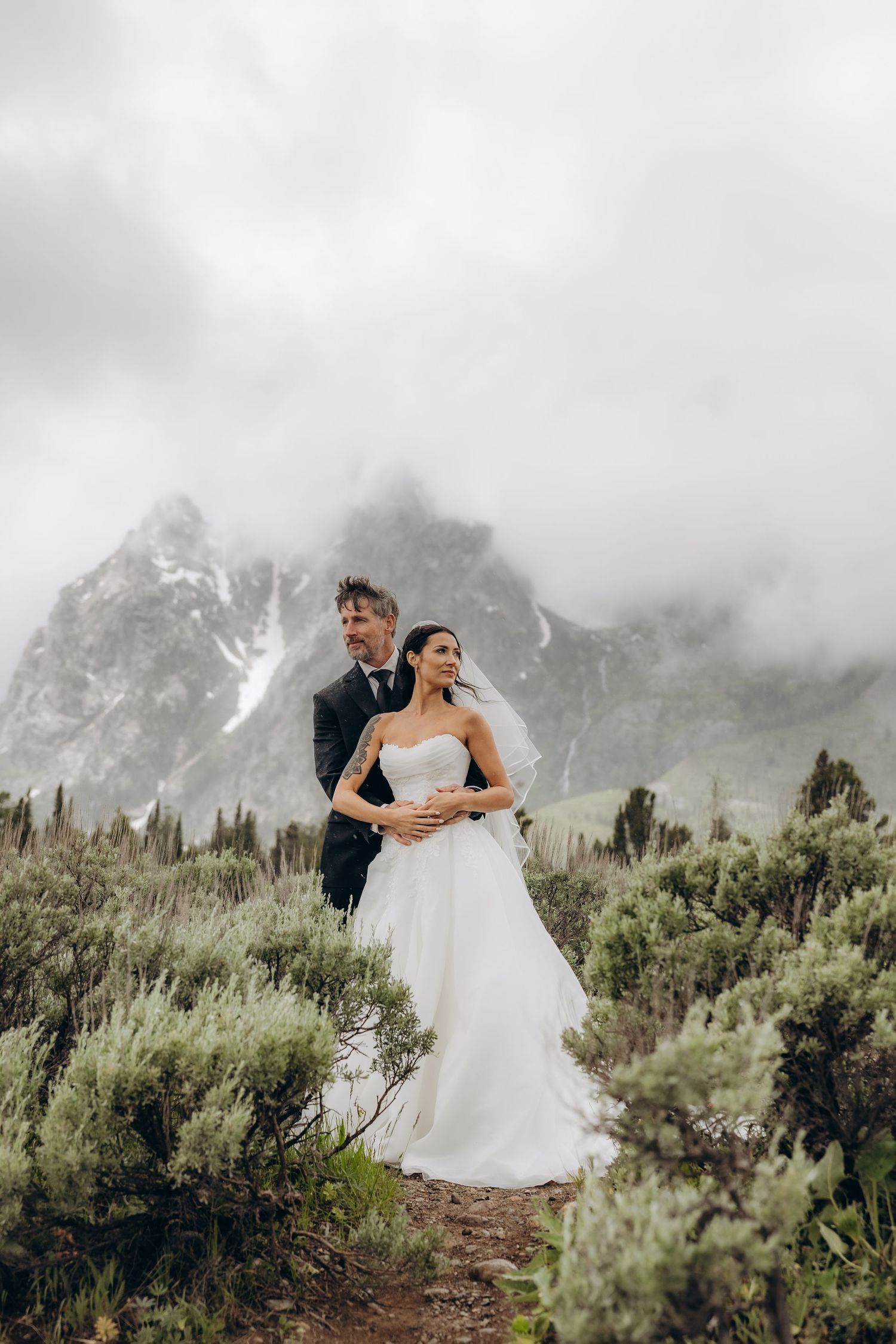 Bride and groom embrace in white wedding attire amid misty mountain landscape with lush greenery and dramatic cloudy peaks.