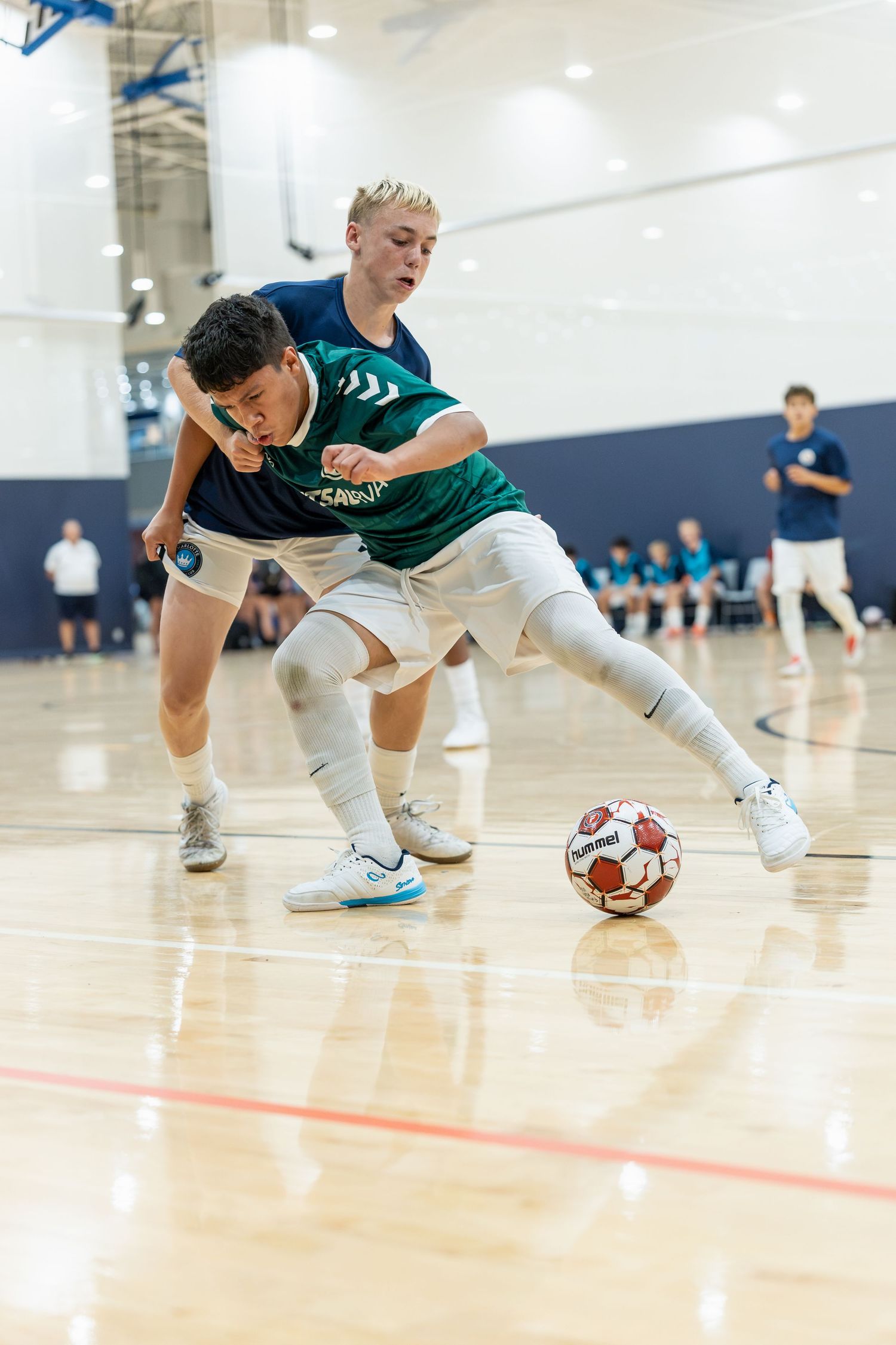 Two youth players compete for the ball during an indoor futsal match on a hardwood court.