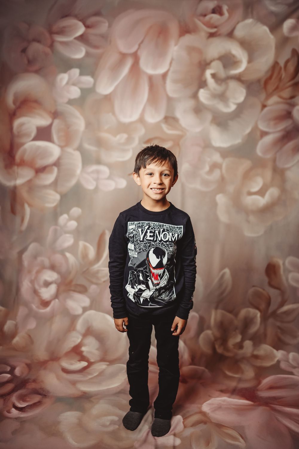 Young boy in a Venom Marvel shirt smiling against a floral painted backdrop in a studio portrait.