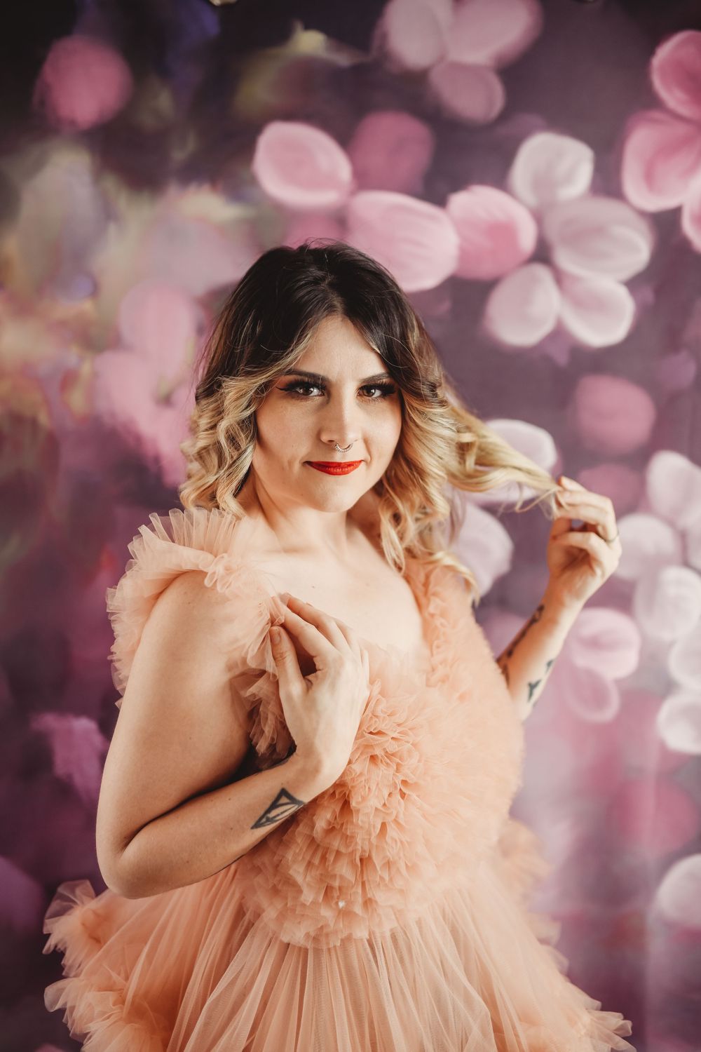 Woman in peach ruffle dress poses against floral pink bokeh background with elegant makeup and blonde highlights.
