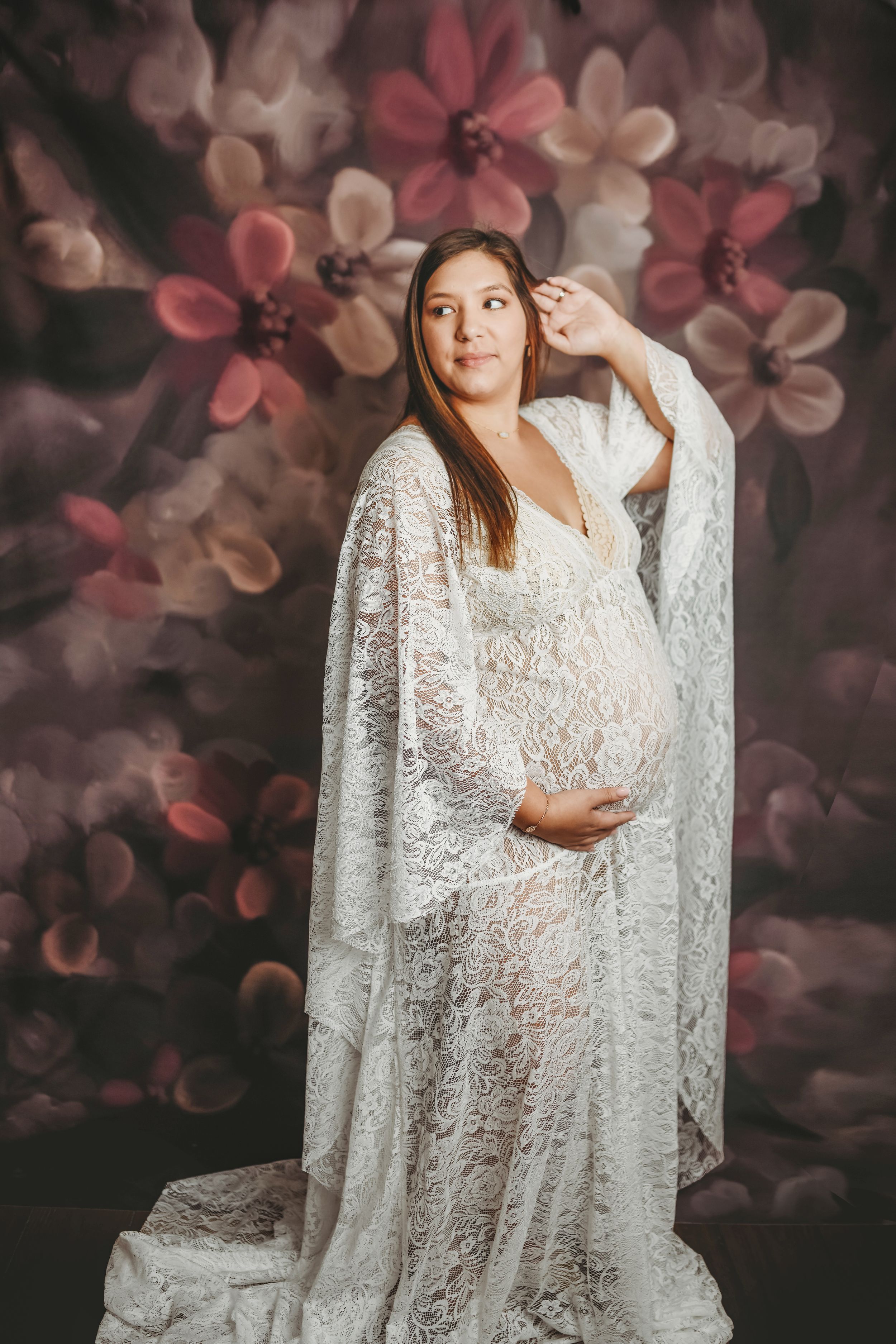 Pregnant woman in white lace gown posing gracefully against a floral backdrop in a maternity portrait.