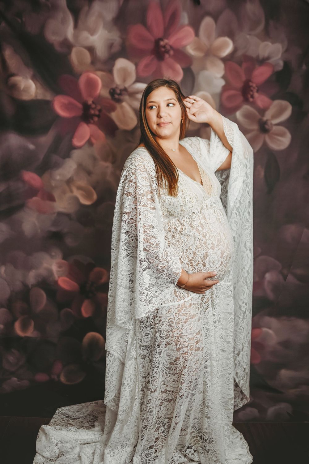Pregnant woman in white lace gown posing gracefully against a floral backdrop in a maternity portrait.