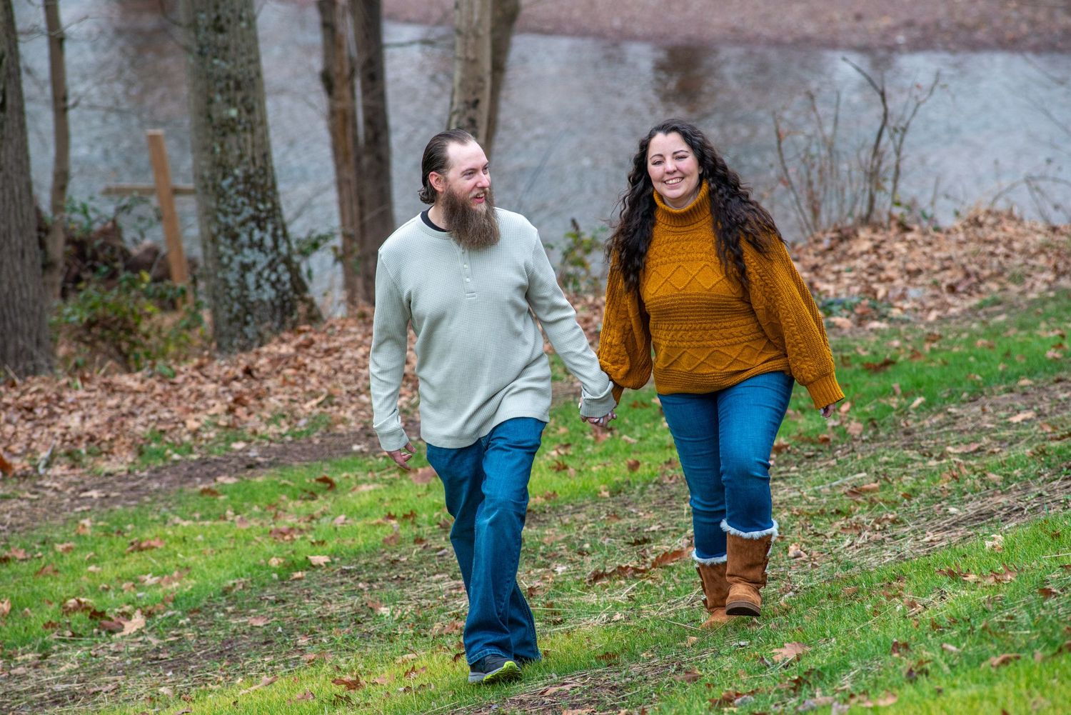 Couple holding hands walking outdoors near a pond in autumn, man with beard in green sweater, woman in mustard sweater.