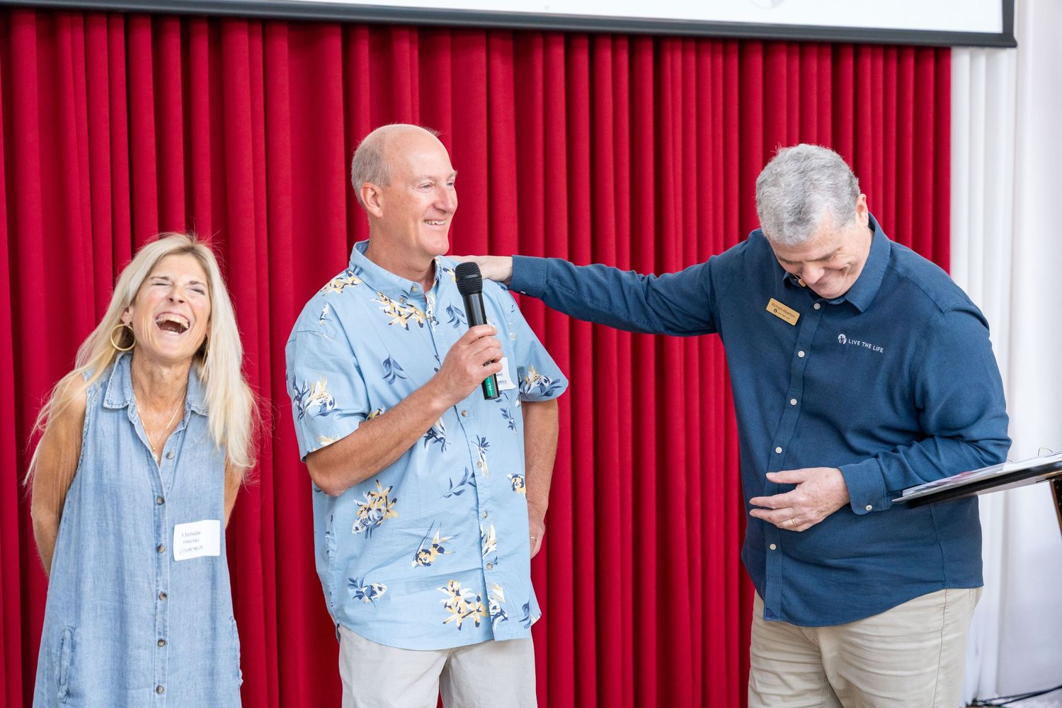 Three people laughing on stage in front of red curtain, man holding microphone at casual event.