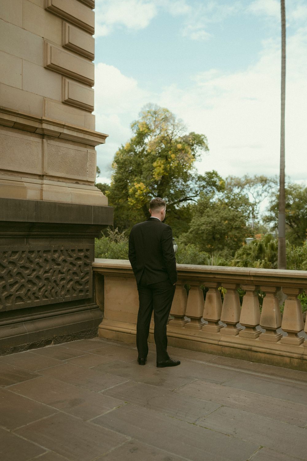 Groom in black tuxedo standing on stone balcony of elegant heritage building overlooking lush green gardens.