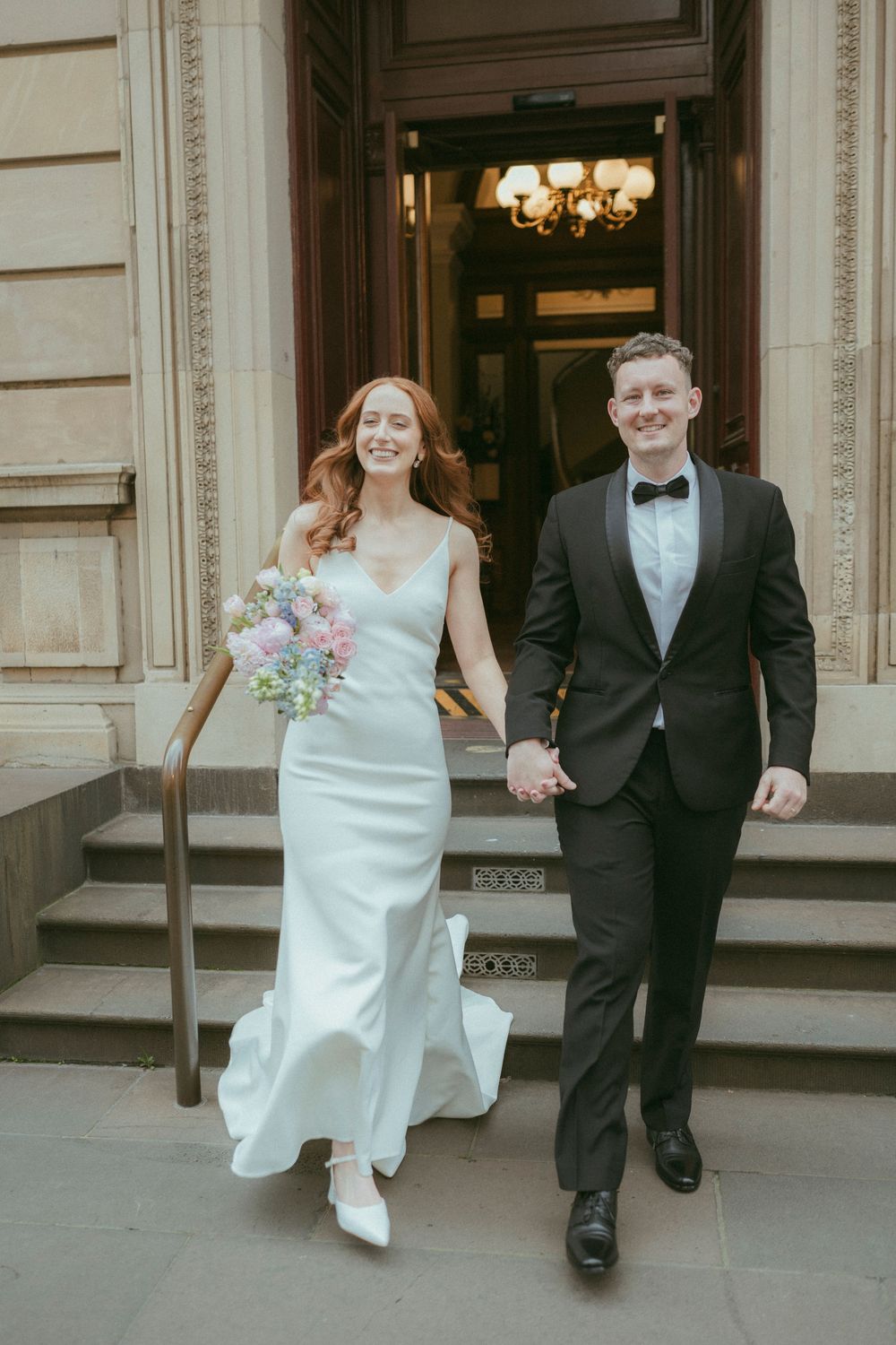 Smiling newlyweds hold hands descending stone steps, bride holding a pastel bouquet in a sleek gown.