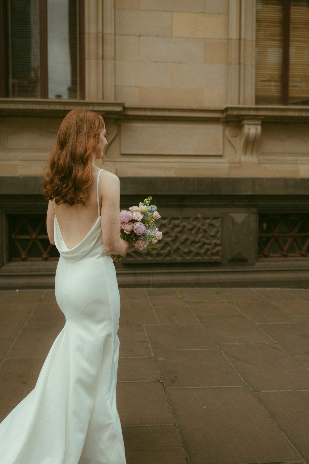 Bride in white backless satin gown holding pink bouquet, standing on stone pavement outside historic building.