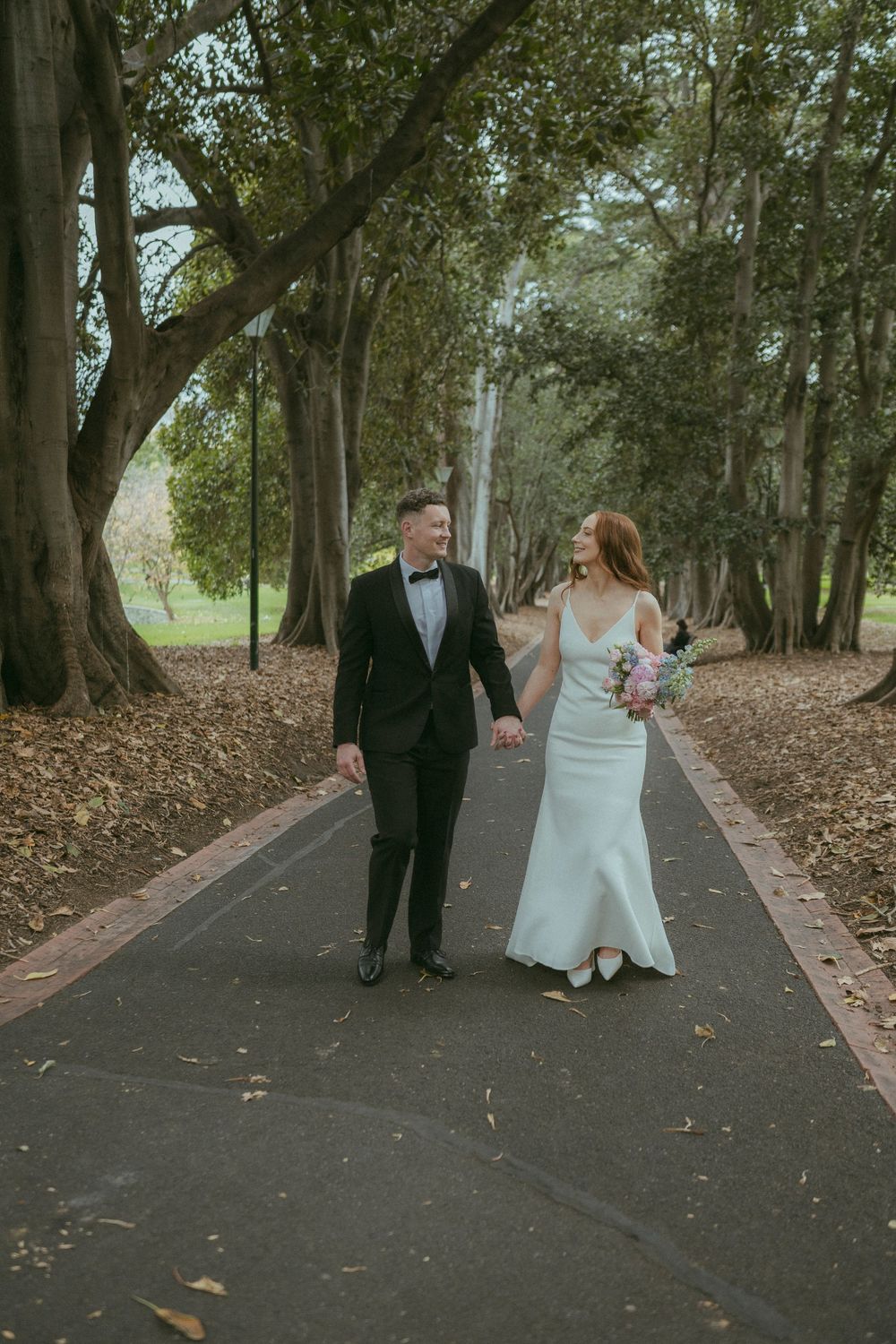 Bride and groom walking hand in hand along tree-lined path, bride holding pastel bouquet in white gown.