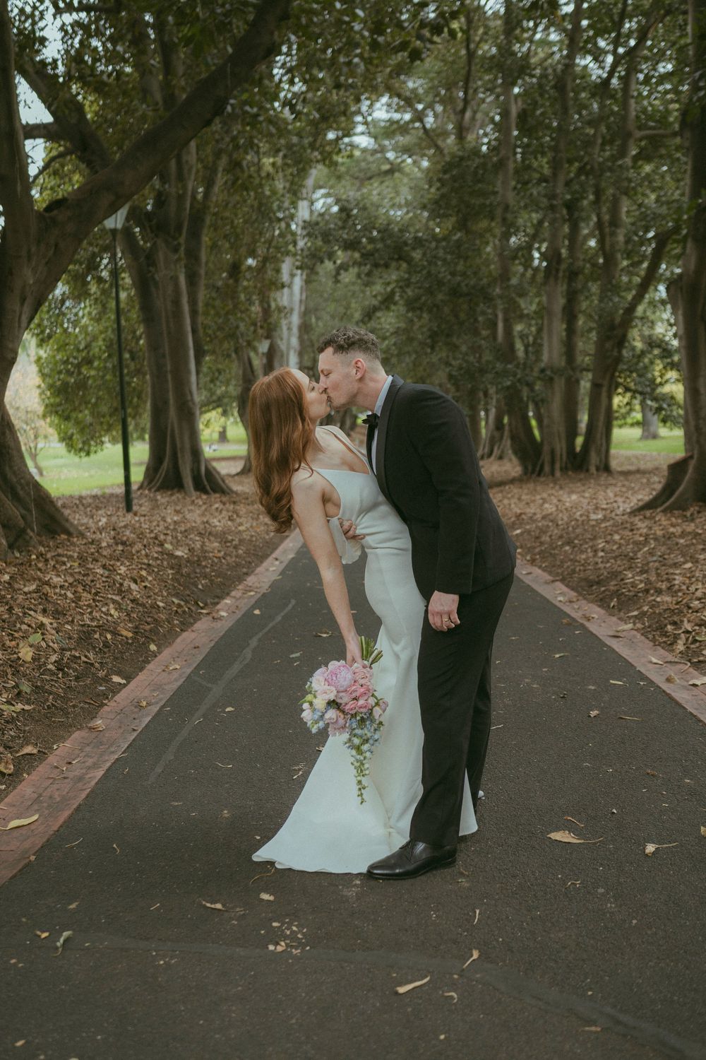 Bride and groom share a romantic kiss on a tree-lined path, bride holding a pink and white bouquet.