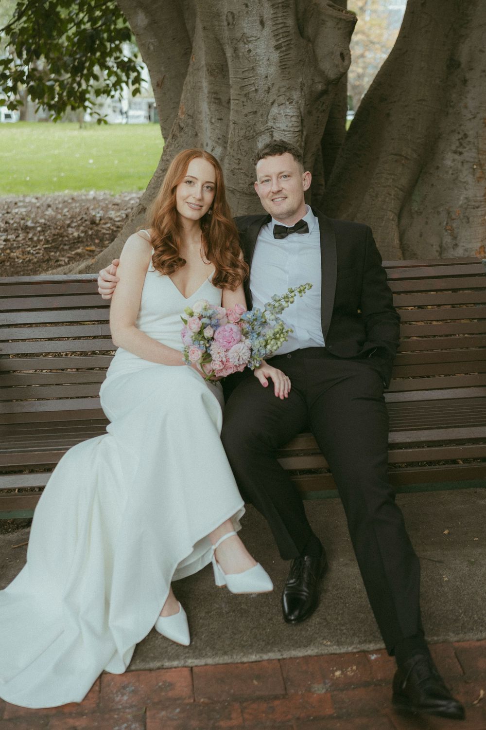 Wedding couple sits on a park bench under a large tree, bride holding a pastel bouquet in white dress.