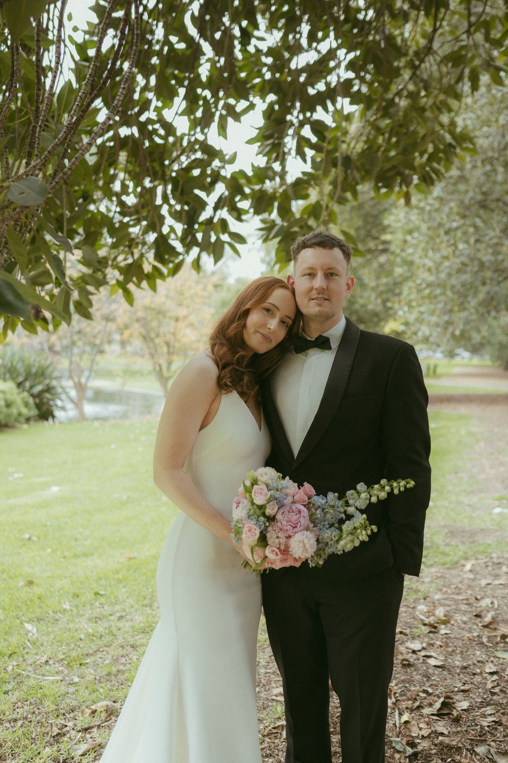 Wedding couple posing under leafy tree, bride in white gown holding pink bouquet, groom in black tuxedo.