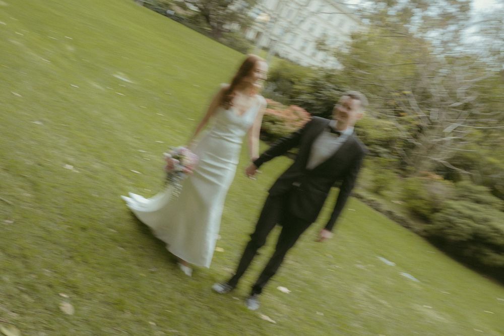 Bride and groom holding hands running joyfully across green park lawn after wedding.