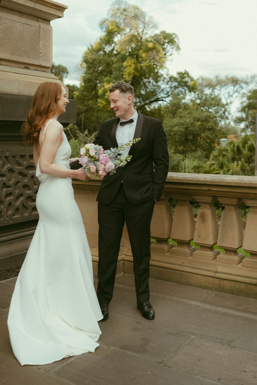 Bride and groom share a romantic moment on a stone balcony, she holding a pink bouquet in a white gown.