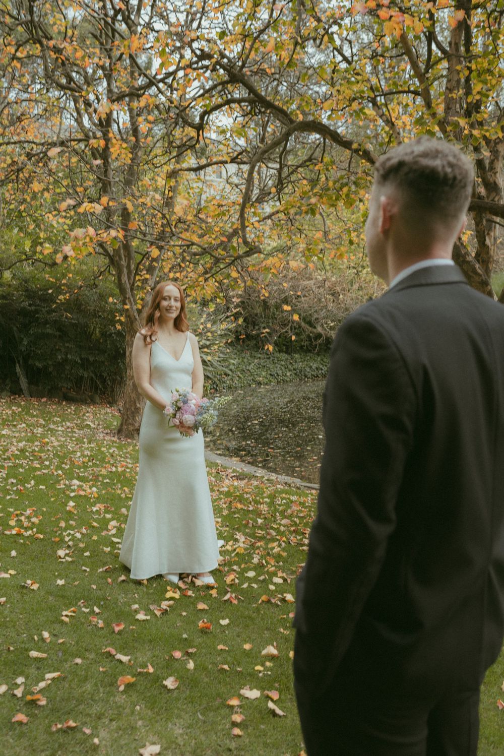 Bride in white gown holds bouquet facing groom in black suit amid autumn foliage in garden setting.