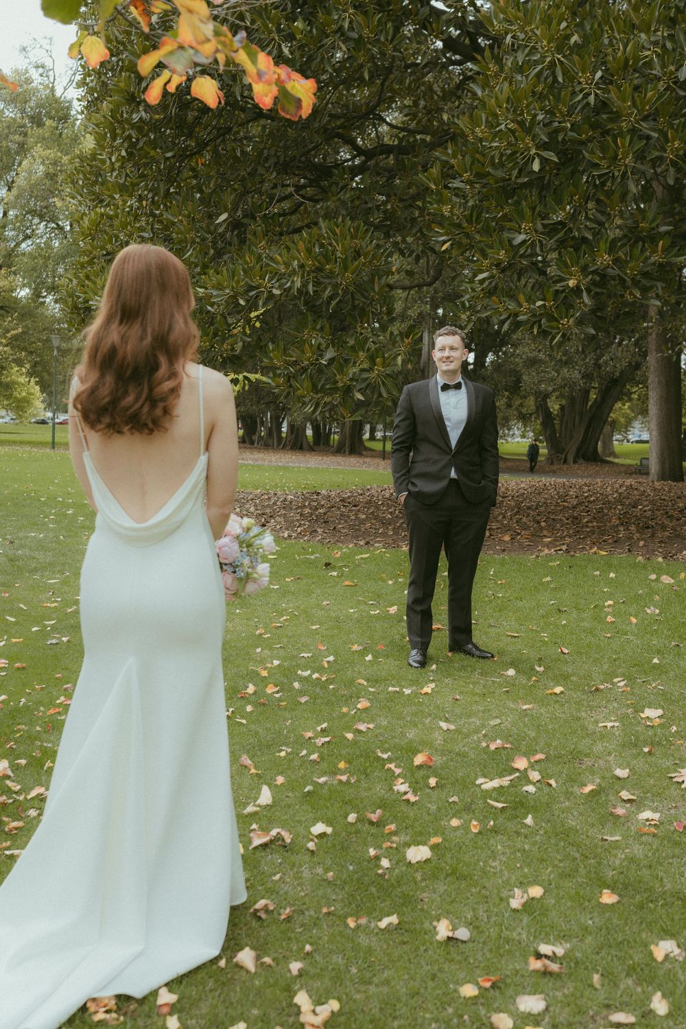 Bride in white backless gown holds bouquet facing groom in black tuxedo in autumn park.