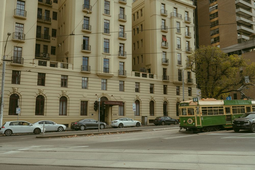A green vintage tram passes a grand cream-colored historic building on a wide city street with parked cars.