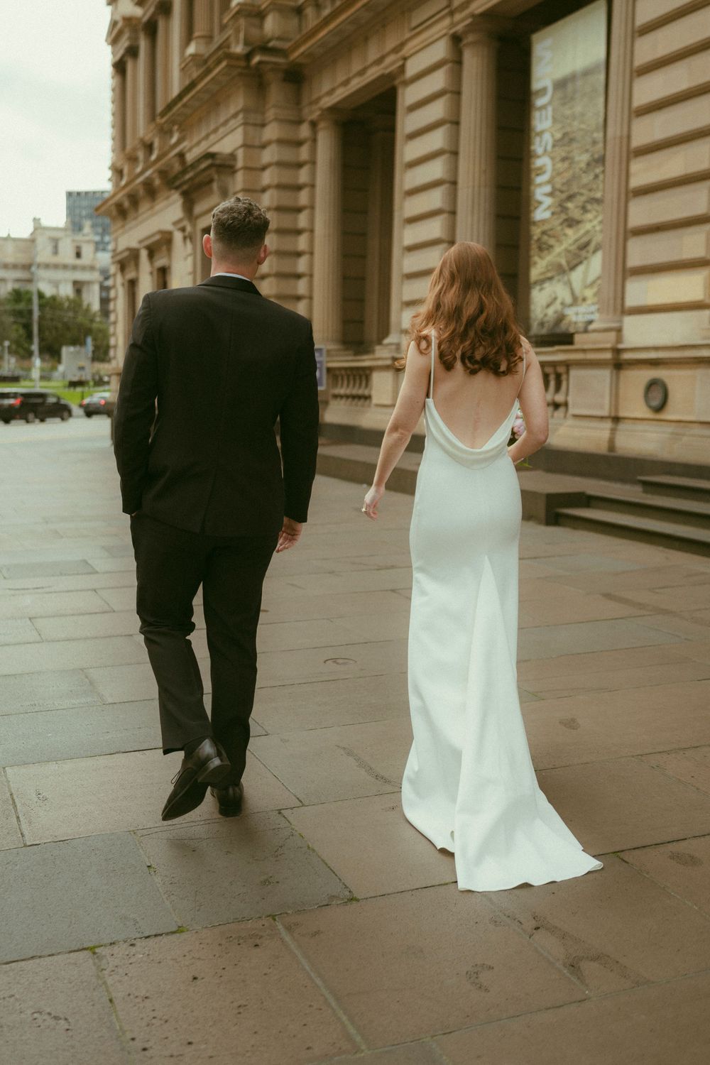 Bride in white backless gown and groom in black suit walk hand in hand past a grand stone building.