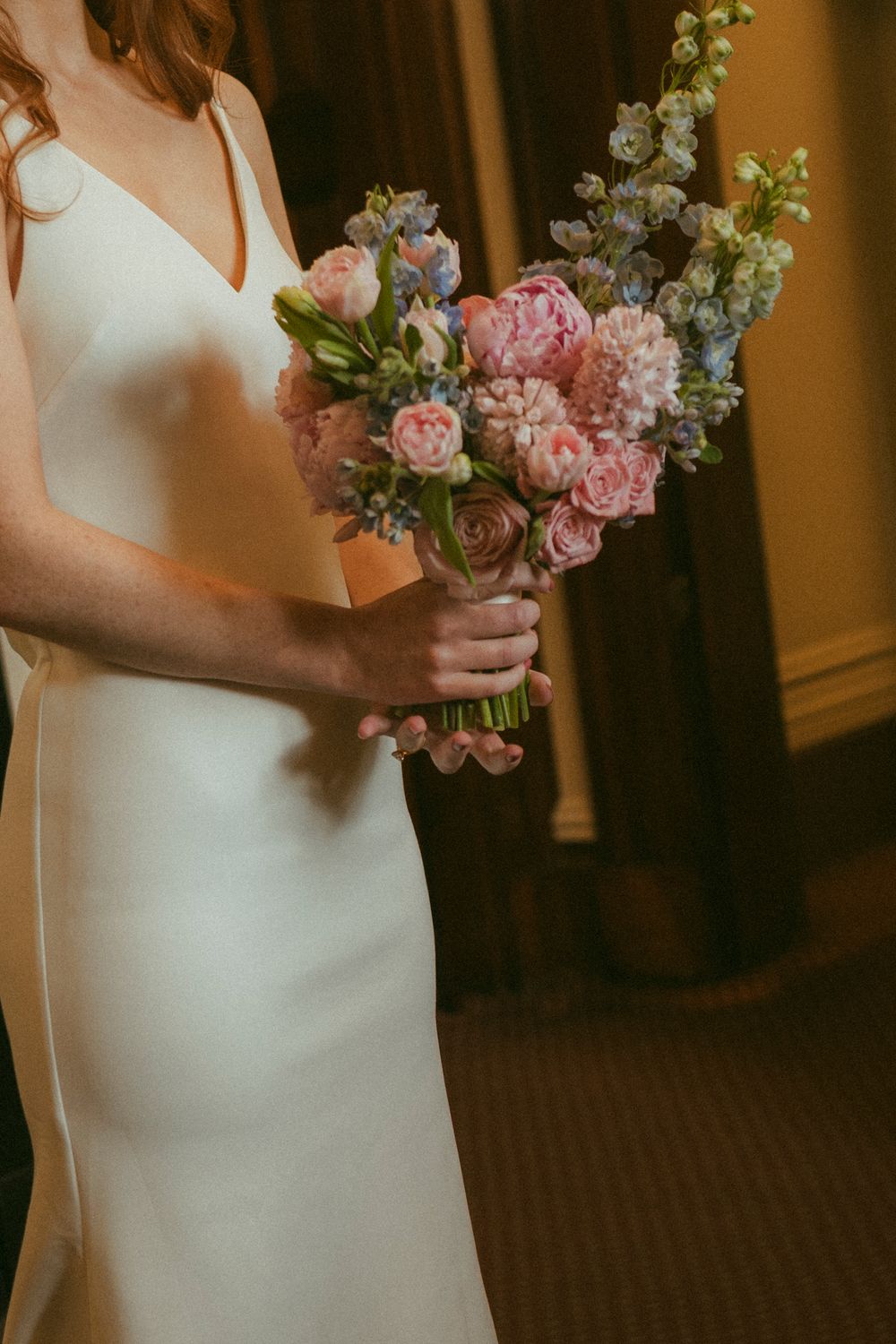 Bride in white gown holding a lush bouquet of pink peonies, roses, and blue delphinium flowers.