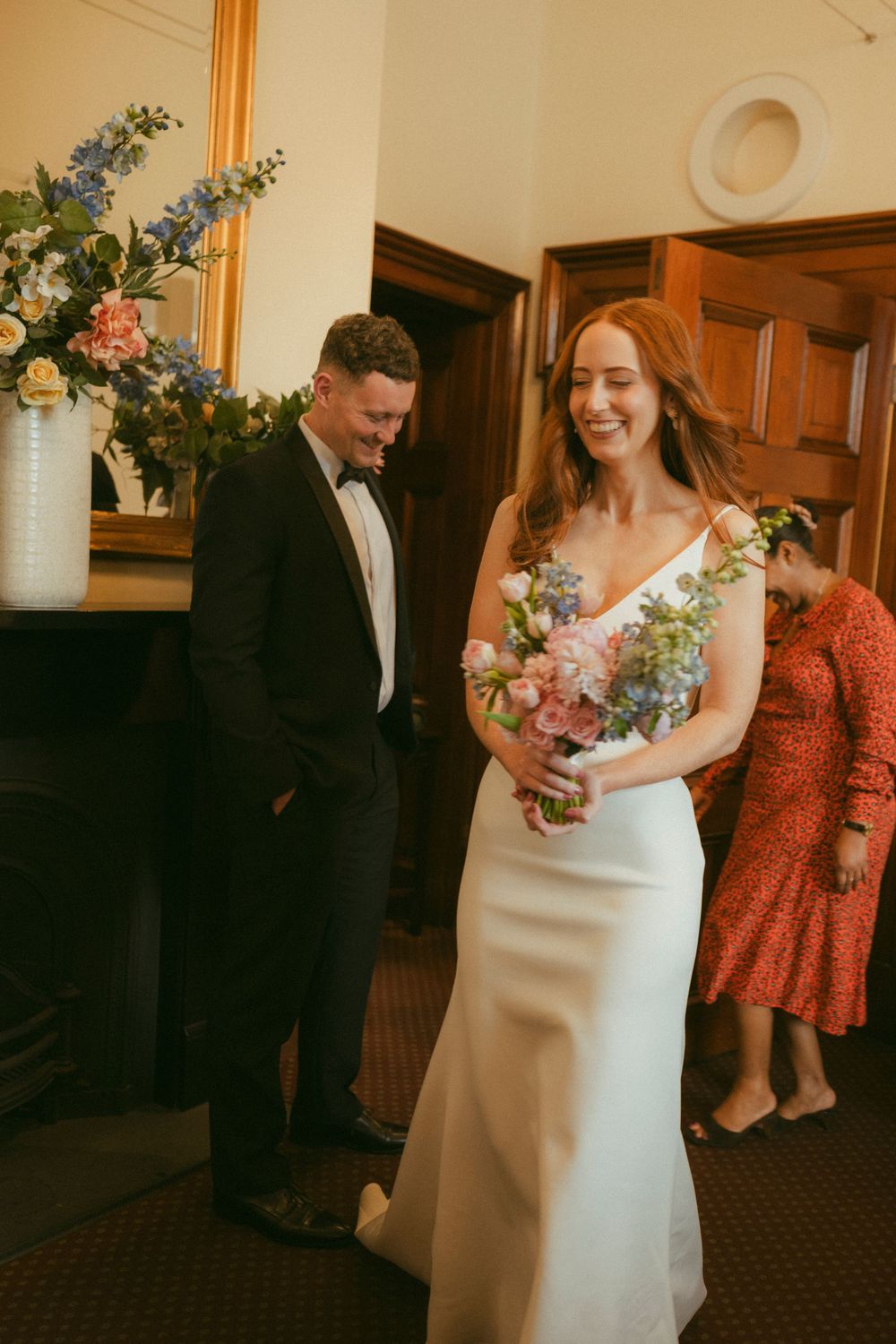 Smiling bride in white gown holds colorful bouquet beside groom in tuxedo at elegant wedding venue.