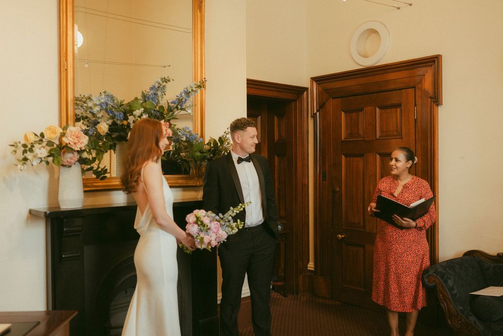 Couple exchanging wedding vows with officiant in elegant room with floral arrangements.