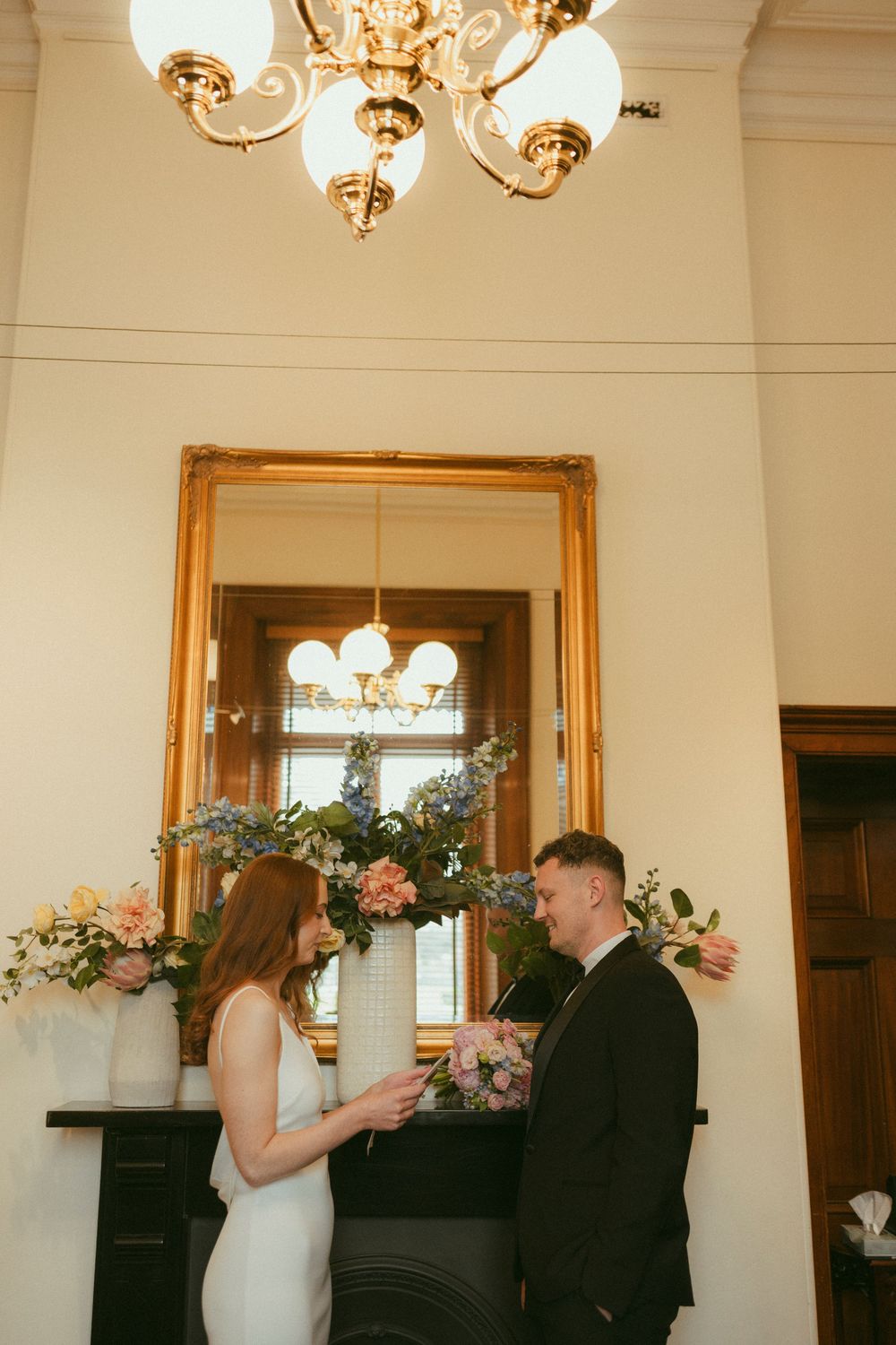 Bride and groom exchange vows indoors by a floral-adorned fireplace with a gold mirror backdrop.
