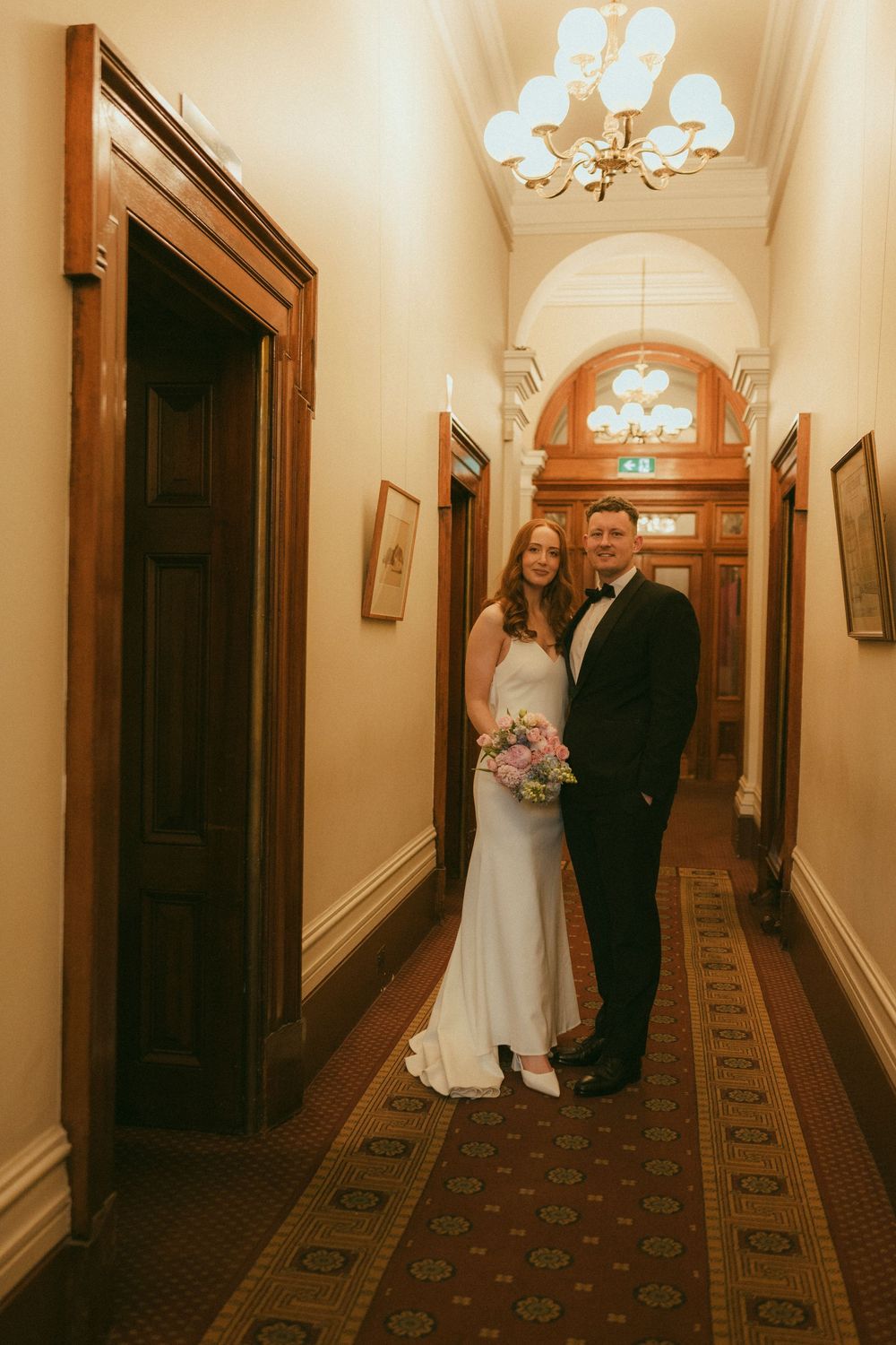 Bride and groom pose in an elegant wood-paneled hallway with chandelier lighting on their wedding day.