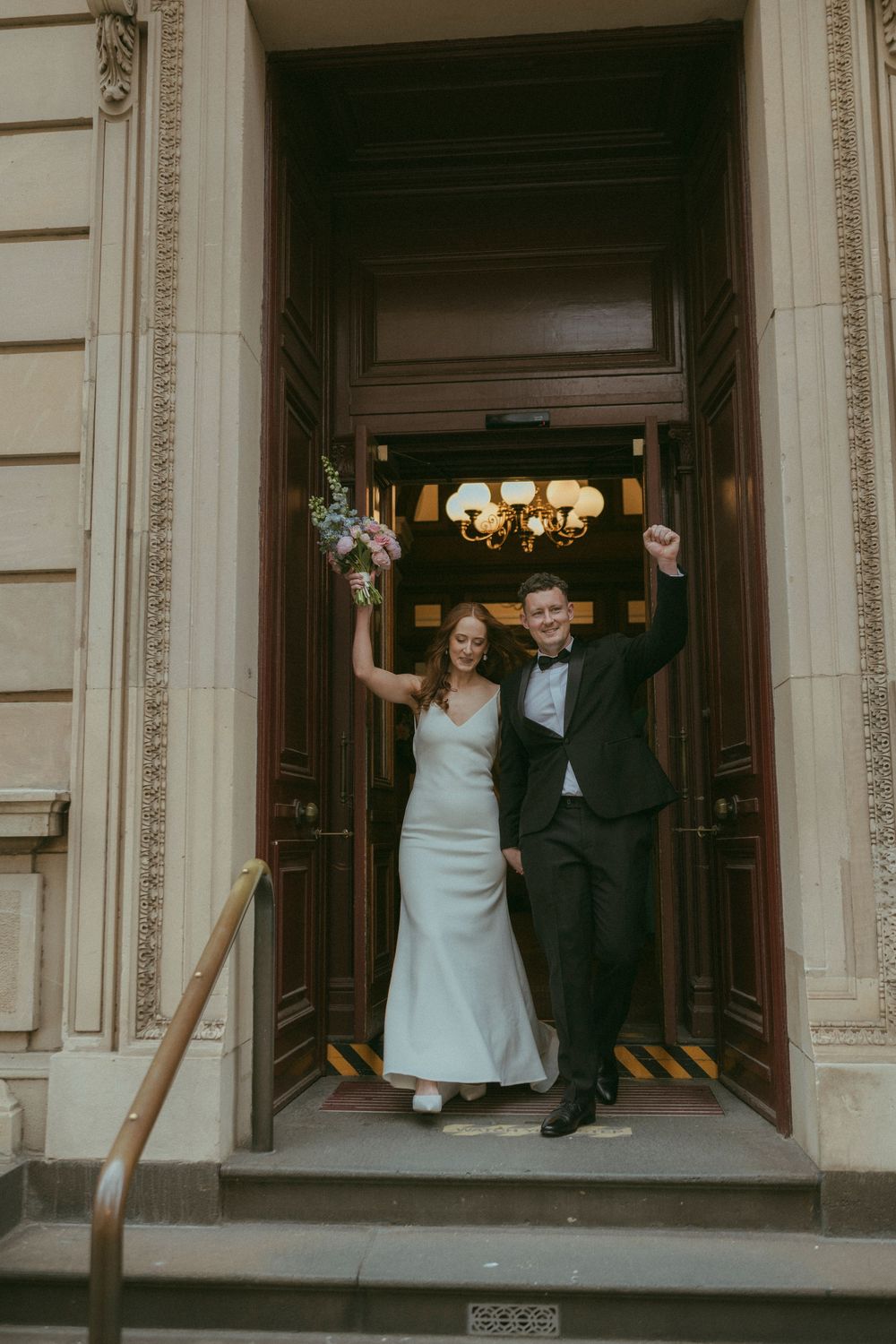Joyful newlyweds celebrate exiting ornate wooden doors of a grand historic building after their ceremony.