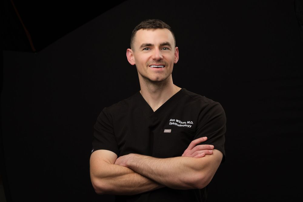 A medical headshot of a physician wearing black scrubs against a black background with his arms crossed.