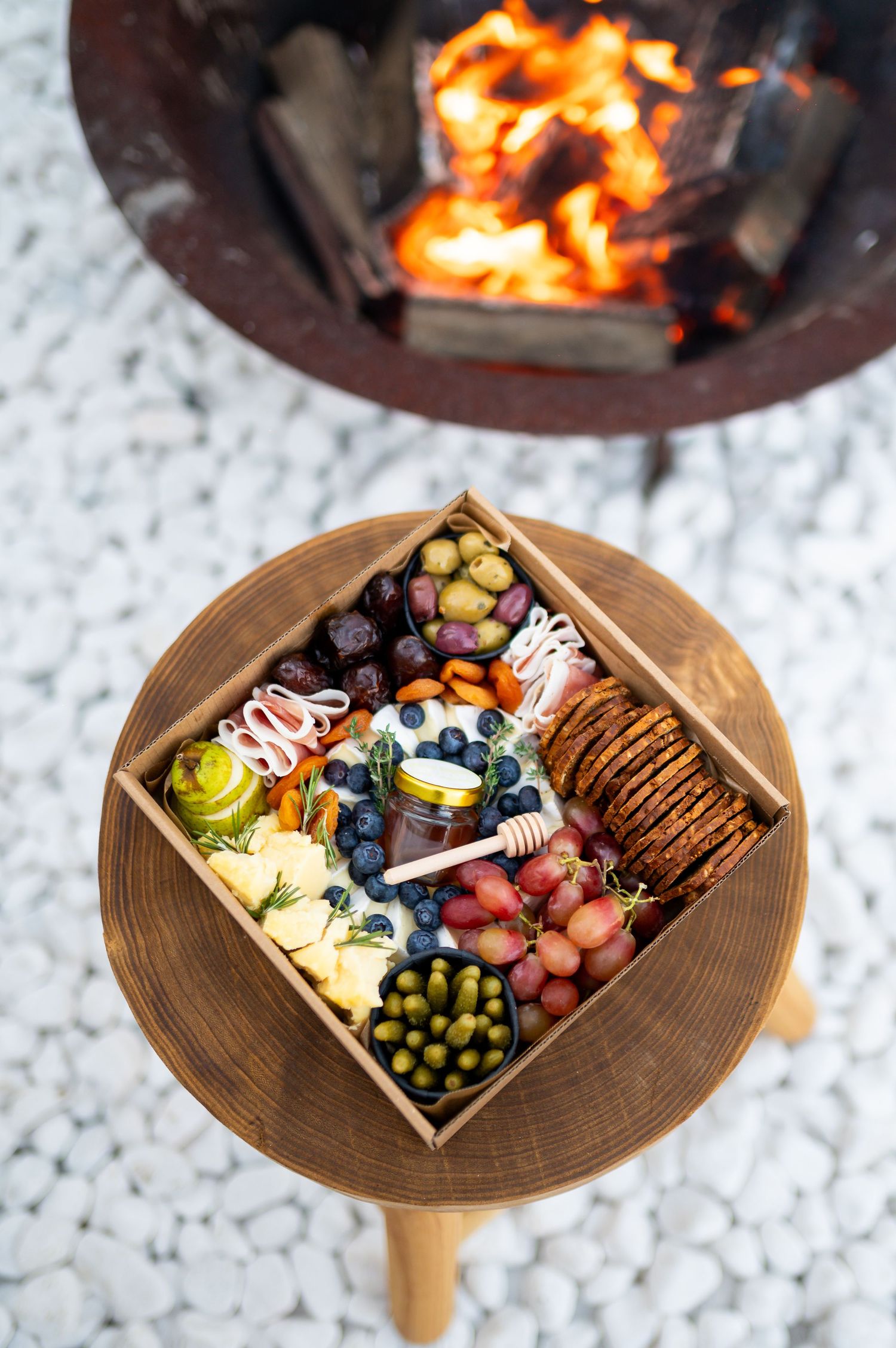 Rustic wooden charcuterie box with grapes, olives, crackers, blueberries, and honey near a glowing fire pit.