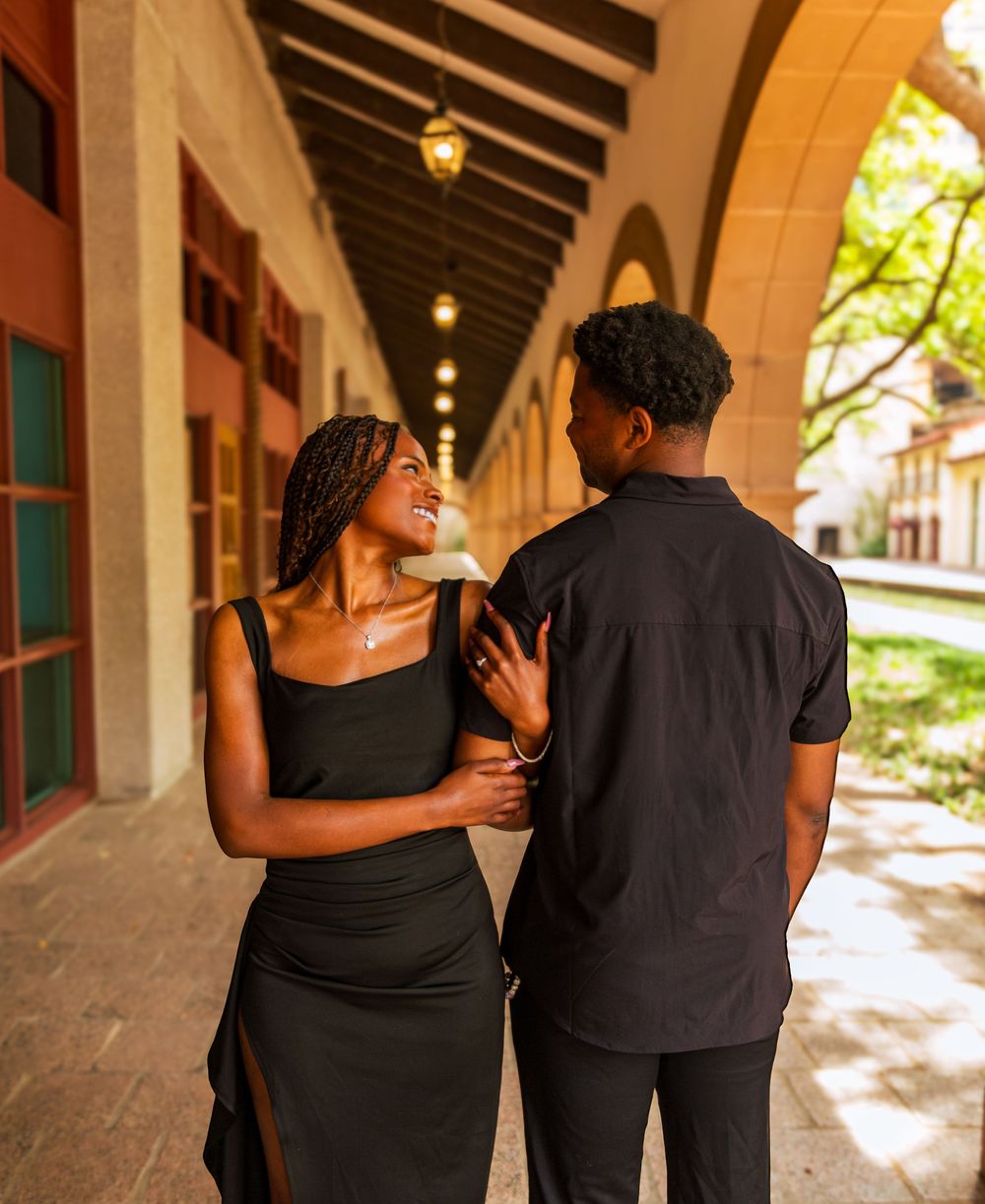 A stylish Black couple in matching black outfits walking together under an arched corridor.