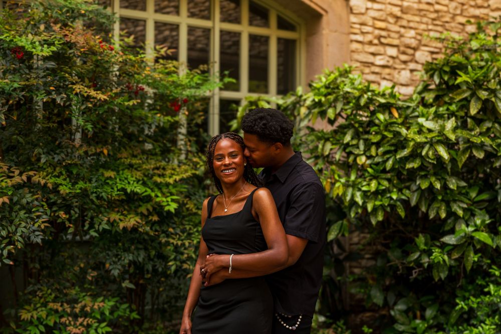 A smiling couple in black outfits embracing in a lush garden with stone wall backdrop.