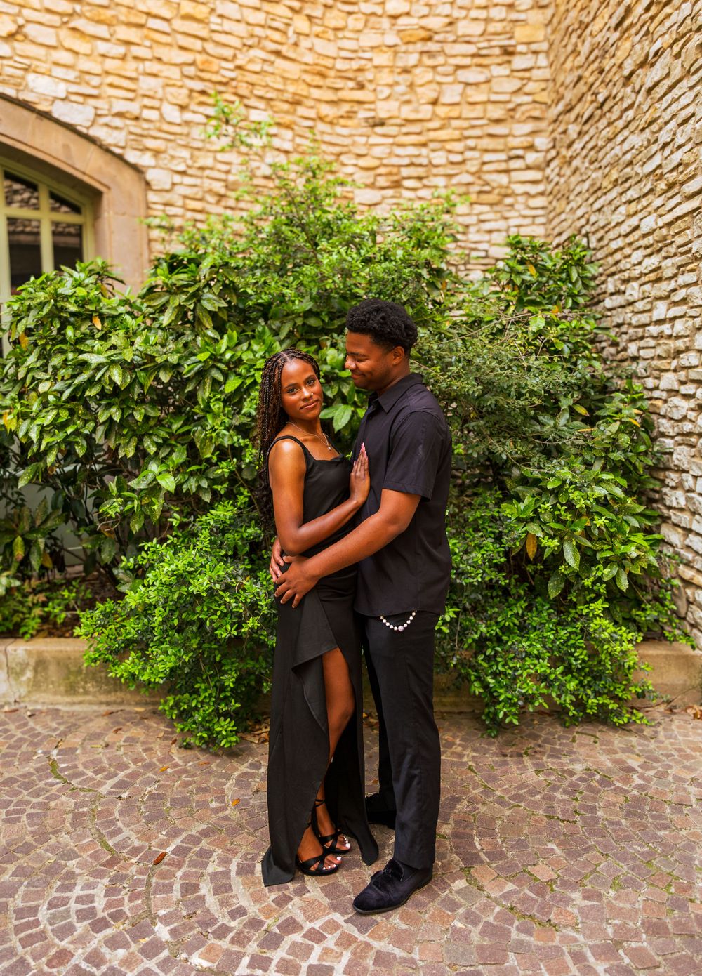 Couple in matching black outfits embracing near stone wall and lush greenery on cobblestone courtyard.