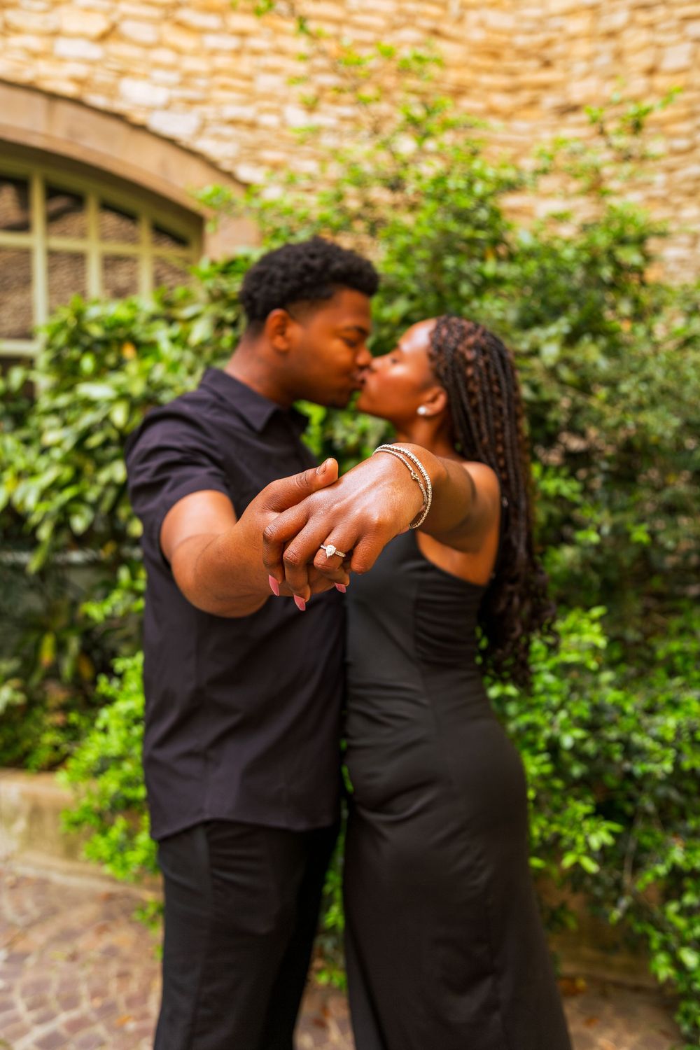 A couple dressed in black shares a kiss while showing off an engagement ring outdoors near a stone wall with lush greenery.
