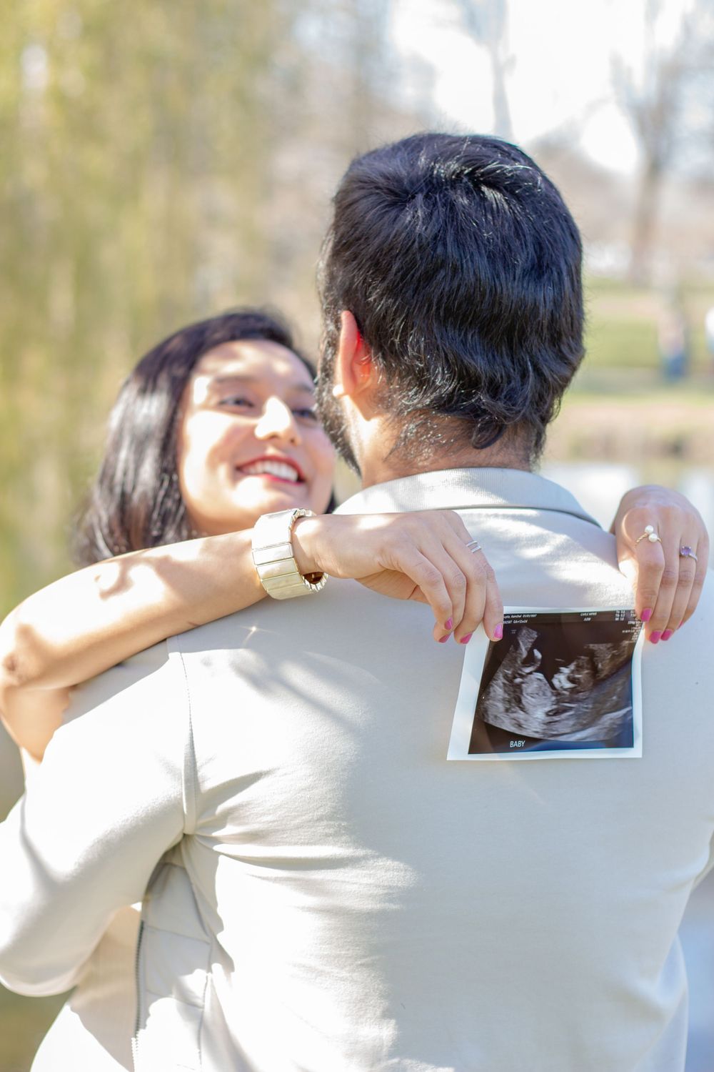 Happy couple embracing outdoors while holding an ultrasound photo, celebrating their pregnancy news in a sunny park.