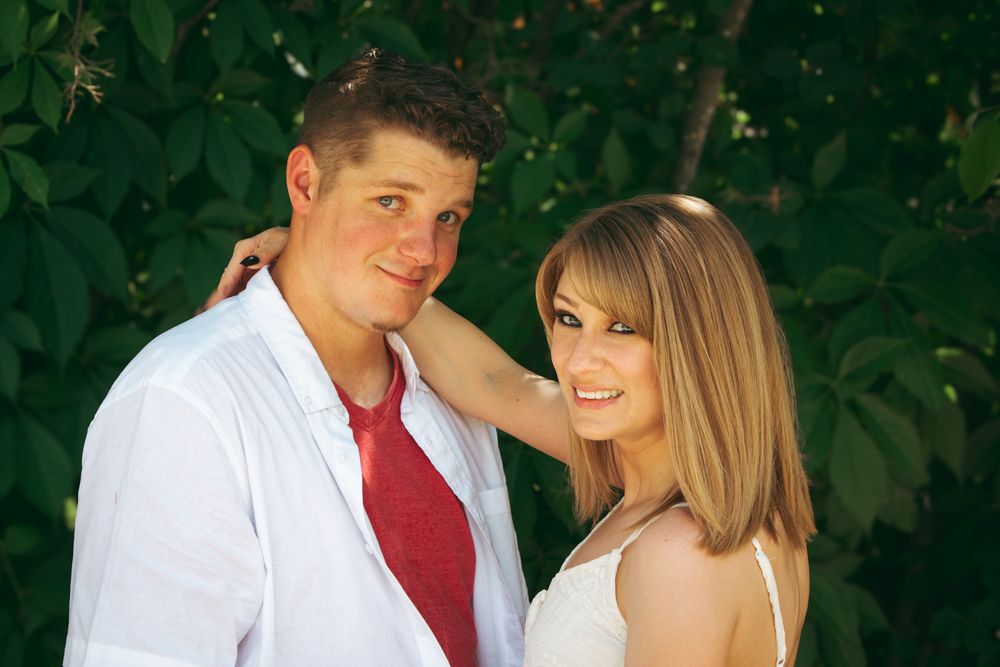 A young couple smiling together outdoors, with green foliage in the background, dressed in semi-formal attire.