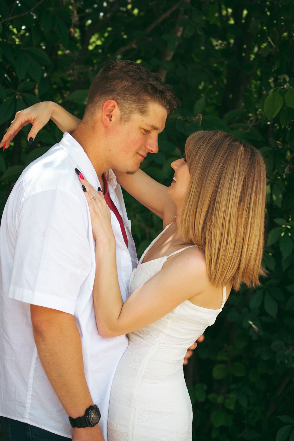 Young couple in white outfits sharing a romantic moment outdoors, surrounded by lush green foliage.