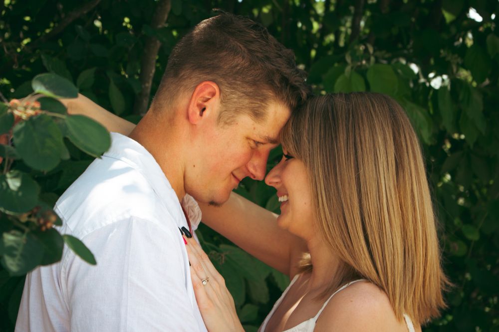 Romantic couple touching foreheads and smiling outdoors, surrounded by lush green trees in a tender moment.