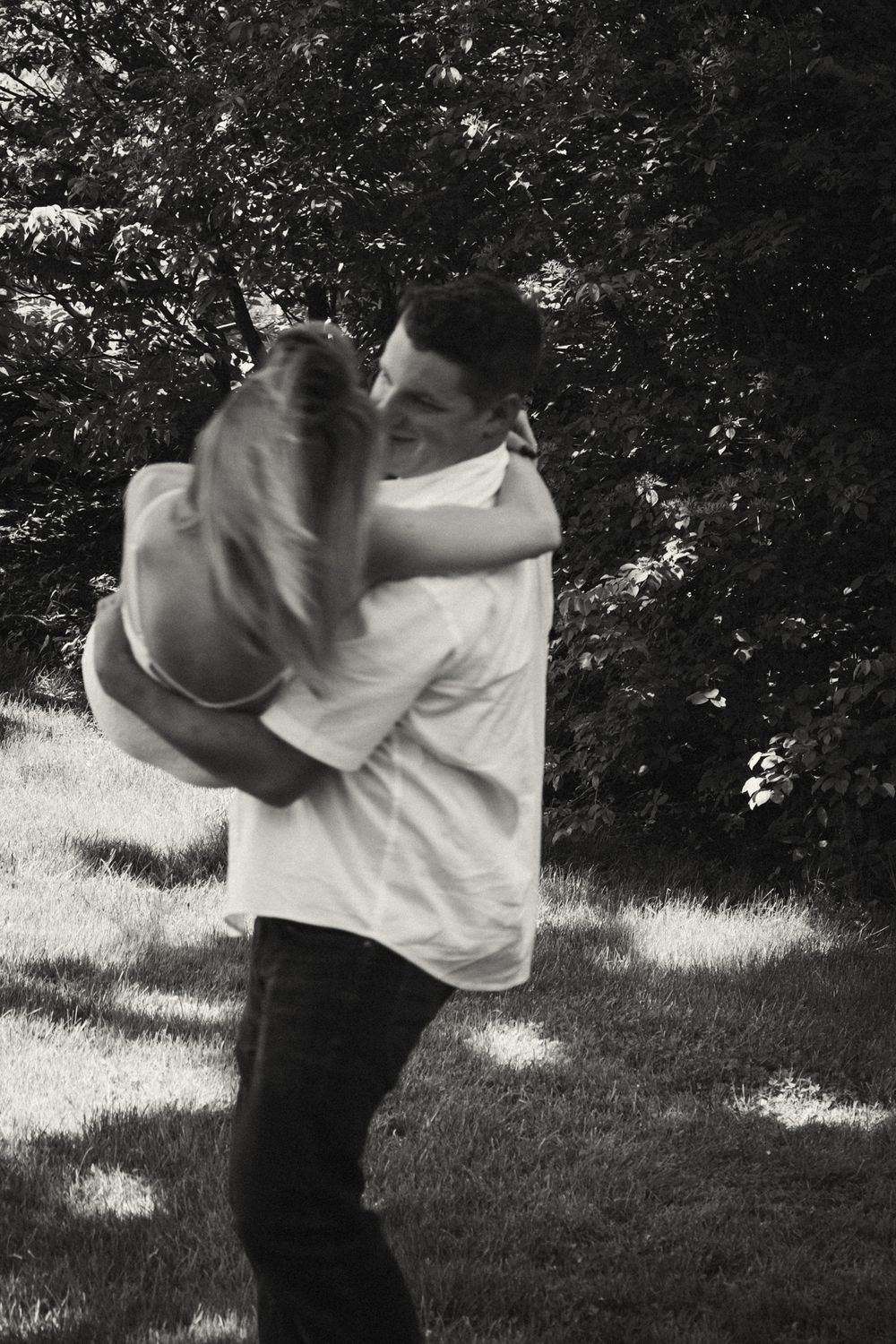 Black and white photo of a couple embracing and dancing outdoors on a sunny lawn with lush green trees in the background.