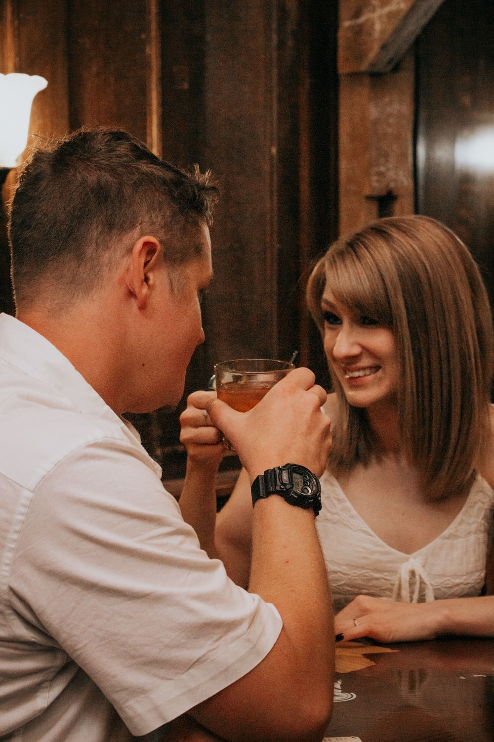 Couple sharing a romantic moment over drinks at a cozy bar, woman smiling warmly at man wearing a black watch.