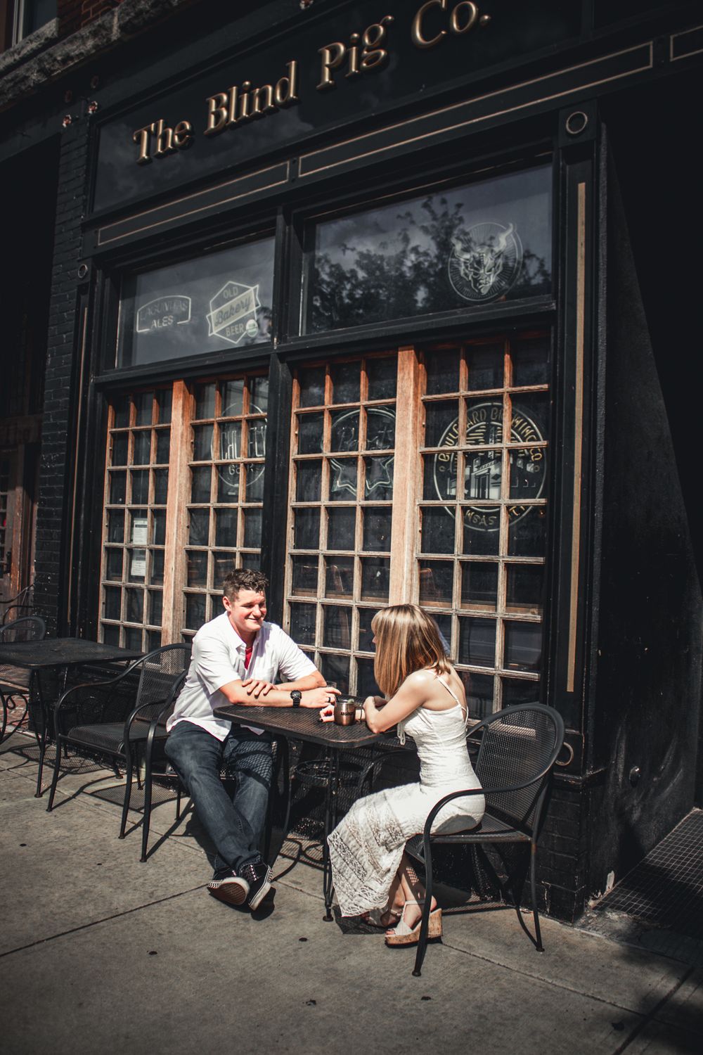 Couple enjoying drinks at outdoor table in front of The Blind Pig Co. bar on a sunny day.