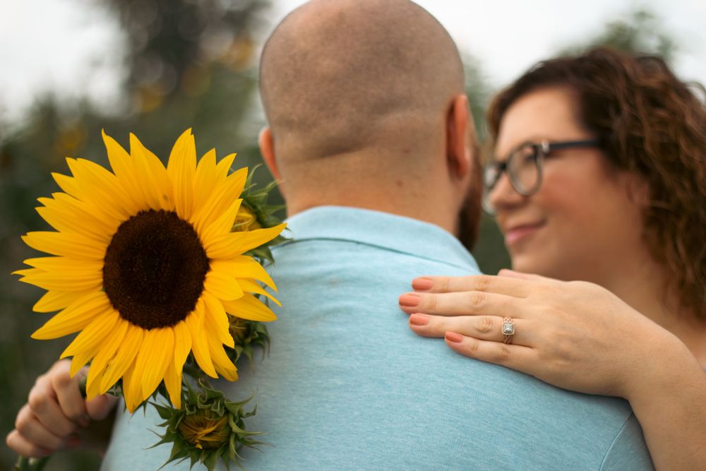 A woman with glasses embraces a bald man holding a bright yellow sunflower outdoors.