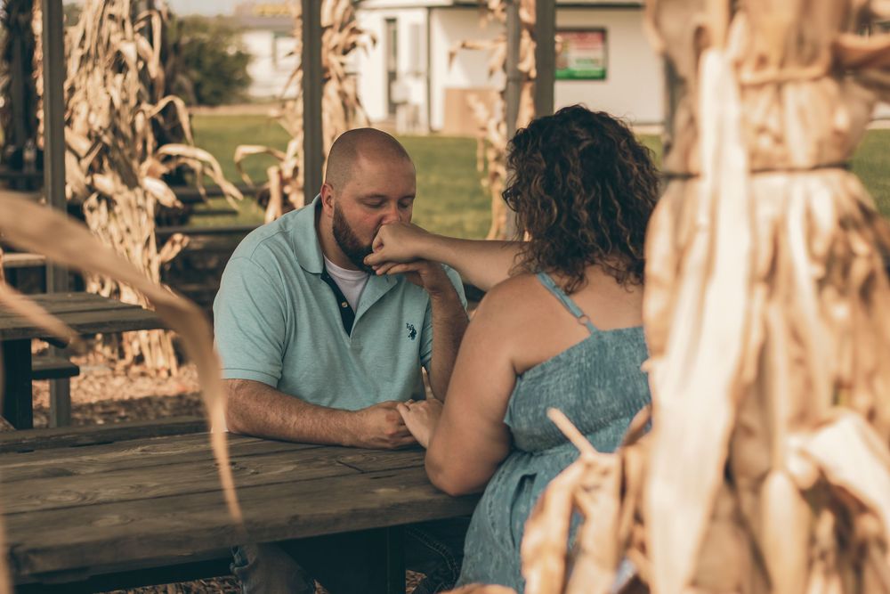 A man in a blue polo shirt kisses a woman's hand at an outdoor picnic table near a cornfield.