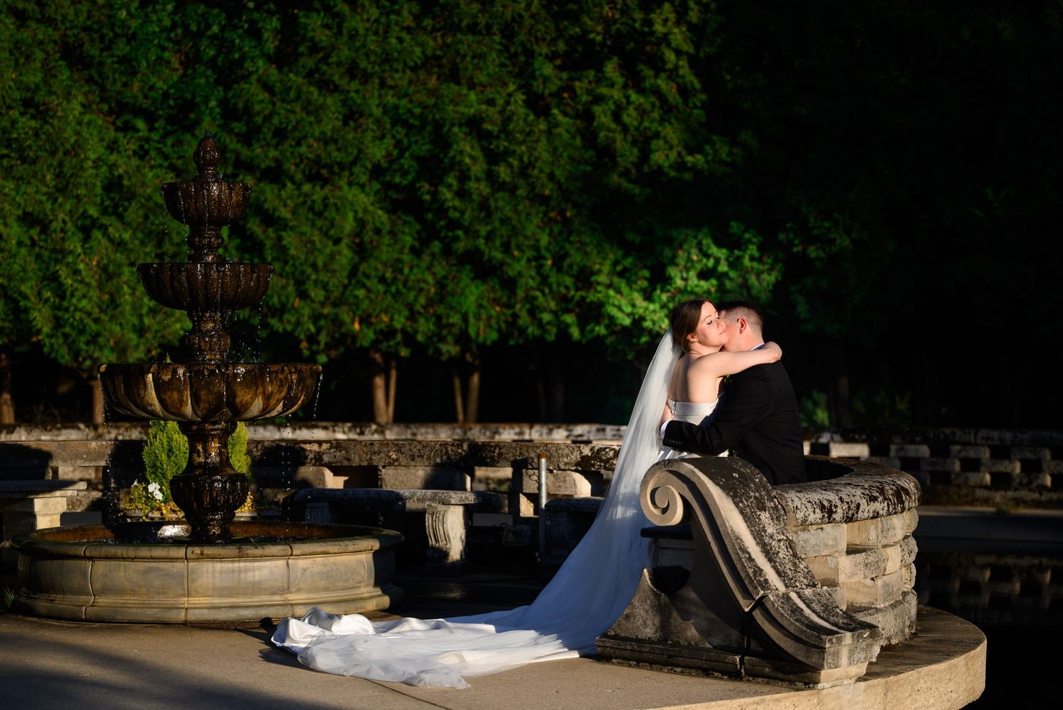 Bride and groom share an intimate moment leaning close together beside a vintage car at dusk outside an elegant brick venue.