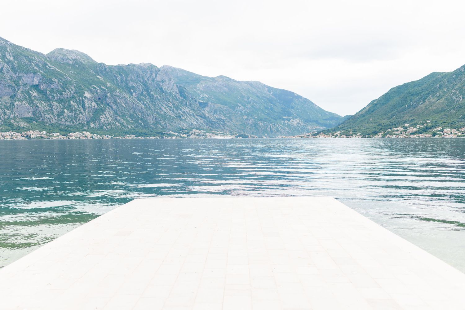 White dock extending into calm bay with clear turquoise water and lush mountain backdrop under overcast sky.
