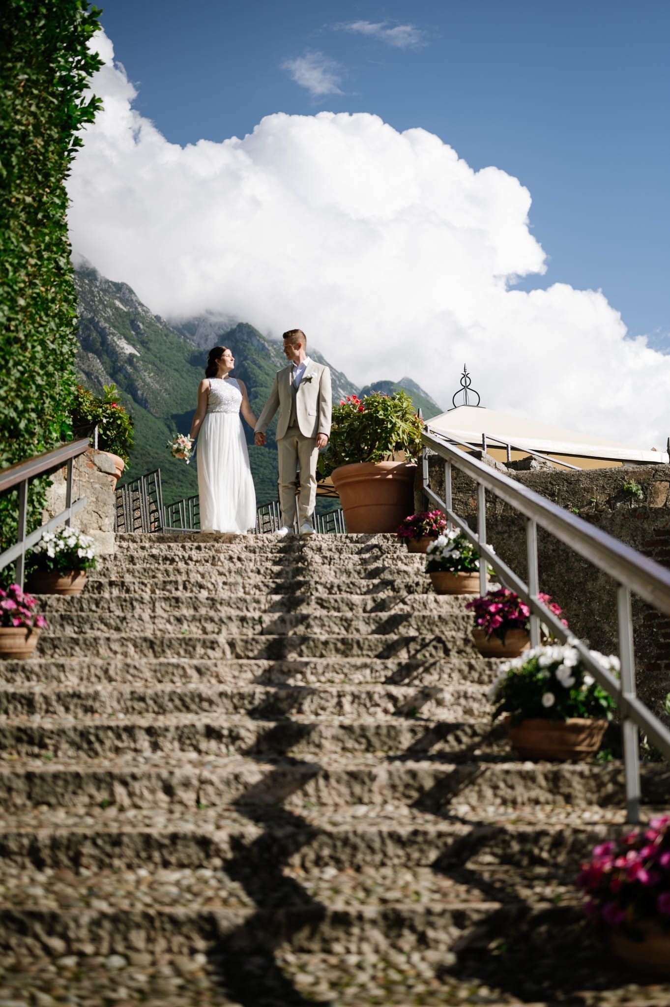 Bride and groom stand atop stone steps adorned with flower pots, with mountains and clouds behind them.